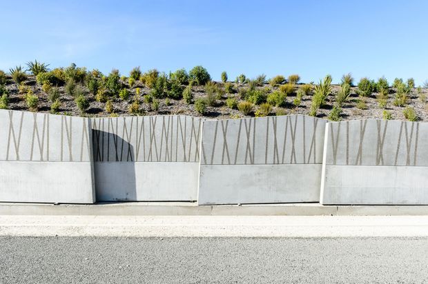 A minimalist streetscape showing a long pale concrete retaining wall with narrow vertical patterned grooves, bordered by a paved road and sidewalk. A landscaped embankment with drought-tolerant plantings rises behind the wall under a clear blue sky.
