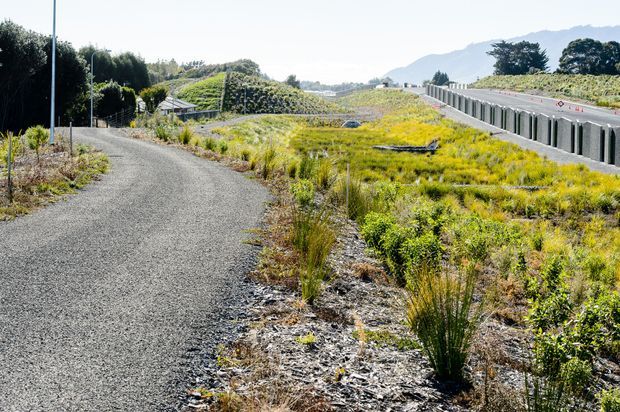 A landscaped outdoor pathway of dark gravel curves through low native-style planting and grassy vegetation. A modern elevated roadway or bridge runs along the right side, with distant hills and trees beyond. The image primarily documents the designed landscape and access path rather than a building.