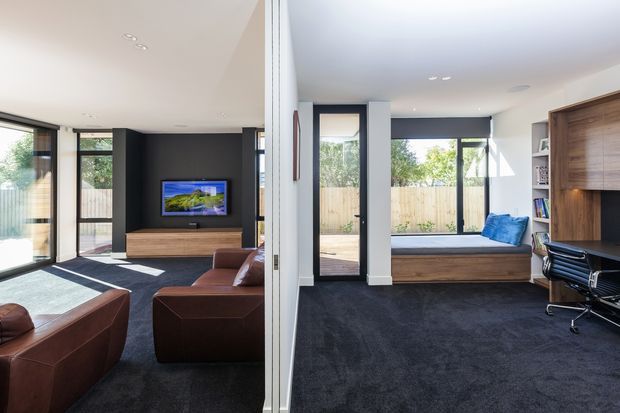 Bright contemporary interior showing an open-plan living room and home office separated by a central wall. The living area has dark carpet, brown leather seating, a timber media console, dark feature wall and large glazed doors. The office includes a built-in timber desk and shelving, a window seat with blue cushion, dark carpet and extensive glazing overlooking a fenced outdoor area.