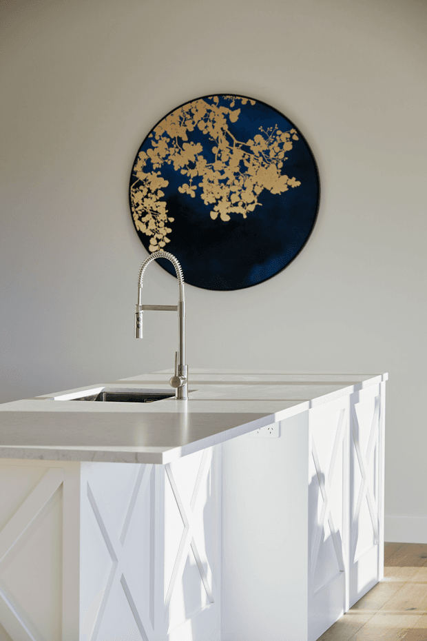 A bright, refined kitchen island with a white geometric feature-panelled base and pale stone benchtop. A stainless-steel gooseneck tap and integrated sink sit beneath a large round navy-blue wall artwork with gold abstract detailing. Light timber flooring and a soft neutral wall create a clean, contemporary setting.