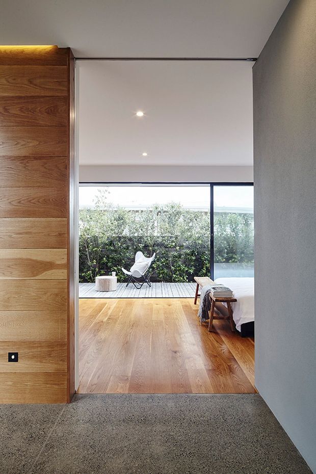View through a wide internal opening into a minimalist bedroom. The room features warm timber wall panelling and matching timber flooring, a low bed partially visible on the right, a small bench, and full-height glazing opening to a timber deck and planted outdoor area. The foreground has a dark textured concrete or terrazzo-like floor, while the bedroom has a clean white ceiling with recessed downlights and simple dark-framed glazing.