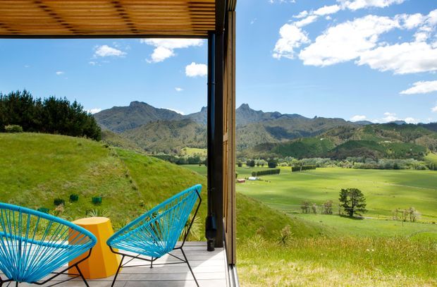 A covered outdoor balcony or terrace viewed from the building, with two blue woven lounge chairs and a small yellow side table on grey decking. A dark vertical structural frame and timber slatted overhead roof provide shelter, opening onto expansive green farmland and rugged mountain ranges under a bright blue sky.