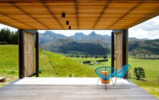 A covered open-air terrace framed by timber-lined ceiling and folding timber shutters, with a broad timber deck and two bright blue outdoor chairs overlooking a lush rural valley and dramatic mountain range. The space has a strong indoor-outdoor connection and expansive landscape outlook.