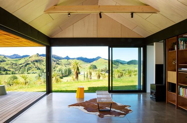 A bright, open-plan living room with full-height black-framed glazing opening onto a deck and expansive green rural hills. The room features a polished concrete floor, a striking timber-lined vaulted ceiling with exposed beam, built-in timber shelving, a low timber lounge chair, a small yellow side table, and a natural hide rug. The glazing provides strong indoor-outdoor connection and panoramic landscape views.