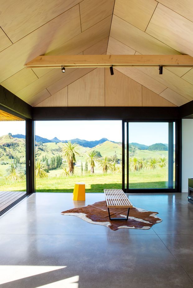 A minimalist open-plan interior with expansive black-framed floor-to-ceiling glazing overlooking a bright rural mountain landscape. The room features a polished concrete floor, a dramatic angular timber-lined vaulted ceiling, exposed dark structural beams, a simple timber slatted bench, a yellow sculptural side table, and a cowhide rug. The space is bright, contemporary, and strongly connected to the landscape.