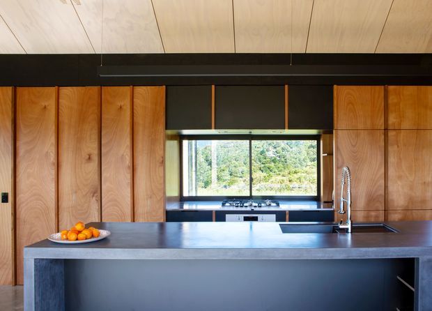 A modern kitchen viewed straight on, centered on a large dark island with an integrated sink and tall gooseneck tap. Floor-to-ceiling timber cabinetry with visible natural grain lines the walls, while a broad window above the rear bench overlooks greenery. The ceiling is clad in pale timber panels, and a dark structural or joinery band creates strong horizontal contrast.