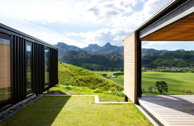 A contemporary architectural view from a landscaped courtyard or terrace between two modern building volumes. The left volume has dark vertical cladding and tall glazing, while the right volume features pale horizontal timber cladding, a deep roof overhang, warm timber soffit, and a large open glazed living area with a timber deck. The composition frames a vivid green rural valley and dramatic mountain ranges beyond.