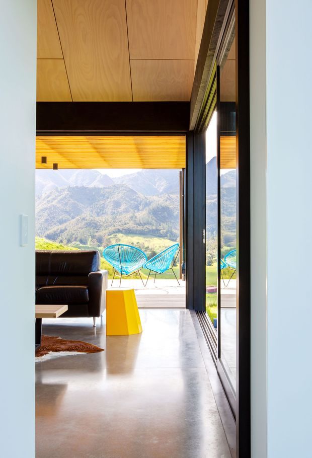 A bright modern living space opening onto a deck with expansive mountain views. The room features a polished concrete floor, dark-framed full-height glazing, warm timber ceiling panels, a black leather sofa, and vivid turquoise outdoor chairs with a yellow geometric side table. Strong indoor-outdoor connection and reflective glazing create a spacious, contemporary atmosphere.