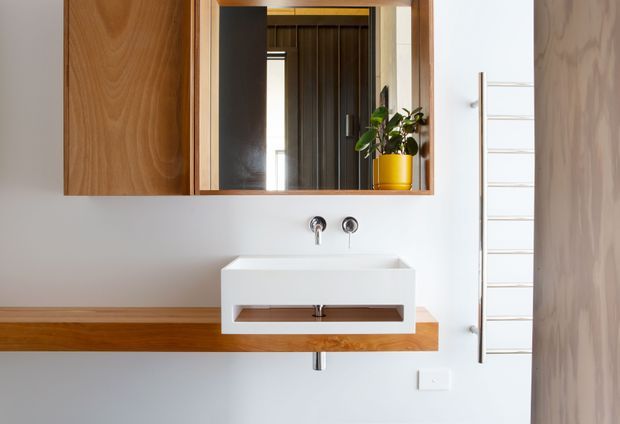 A bright, minimalist bathroom vanity with a wall-mounted white basin, exposed chrome tapware, and a timber drawer shelf. A large timber-framed mirror with integrated upper joinery reflects a dark timber-lined hallway and a potted plant. A slim wall-mounted ladder towel rail is visible to the right, with warm timber detailing contrasting against white walls.