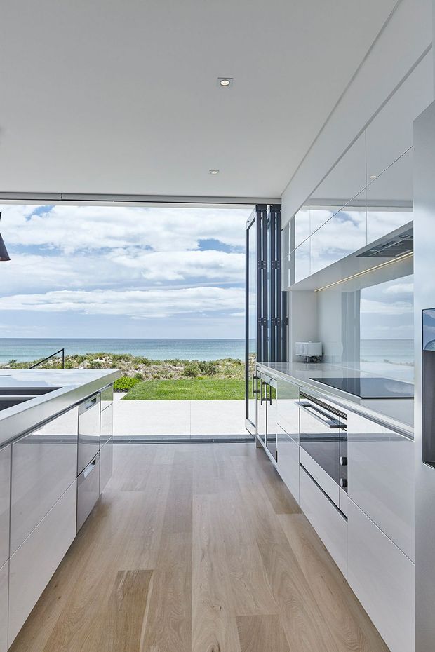 A bright, minimalist contemporary kitchen opening directly onto a landscaped beachfront terrace and ocean view. The room features extensive white handleless cabinetry, integrated appliances, reflective glazing or mirrored cabinetry, pale timber flooring, a white ceiling with recessed downlights, and a large open connection to the outdoors. The scene has a clean, airy, high-end coastal character.