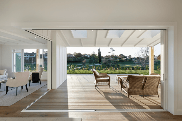 Bright contemporary indoor-outdoor living space with large retractable glazed doors opening onto a timber deck. The room has white walls, exposed white ceiling beams, light timber flooring, and neutral upholstered seating, while the covered terrace contains woven outdoor lounge chairs overlooking a landscaped garden and suburban setting.