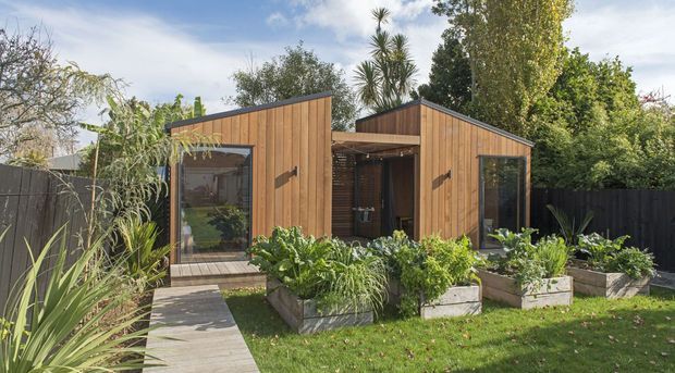 A compact modern timber-clad garden building composed of two linked pitched-roof volumes, with large glazed openings, a central covered entry, and a landscaped garden with raised planter boxes and a timber path. The dark fencing and warm wood create a contemporary backyard studio or small dwelling setting.