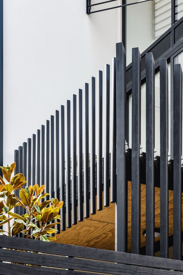 Close-up architectural view of a contemporary stairway or landing enclosed by a series of tall vertical dark timber or metal-look balusters. A dark-framed glazed facade and warm timber flooring are visible behind the balustrade, with ornamental planting at the lower left. The composition emphasizes strong vertical rhythm, contrast between dark elements and white walls, and a modern minimalist character.