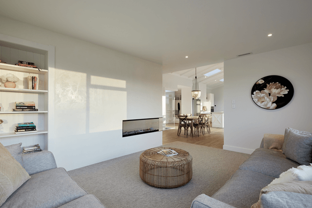 Bright open-plan living room with a large grey sectional sofa, round woven coffee table, built-in shelving, wall-mounted fireplace, and neutral carpet. The space opens to a dining area and kitchen, with pendant lighting, white cabinetry, and strong natural light creating a calm contemporary interior.