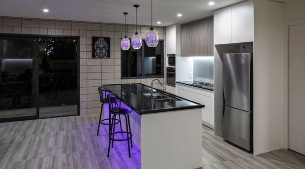 Contemporary open-plan kitchen with a white island, glossy black stone benchtop, integrated cooktop and sink, white and wood-grain cabinetry, stainless-steel refrigerator, and three purple pendant lights. Large dark-framed sliding glazing opens toward an outdoor area, while grey tiled wall cladding and pale timber-look flooring complete the space.