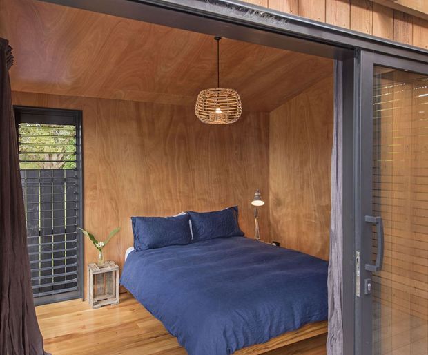 Compact bedroom enclosed by warm timber wall and ceiling panelling, with a timber floor, dark-framed glazed sliding door, louvred window, navy bedding, and a woven pendant light. The room has a cosy, cabin-like character with strong natural wood finishes.