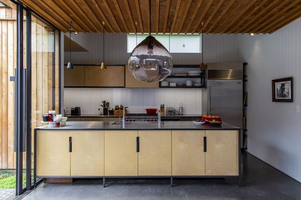 A contemporary kitchen with a large central island, pale wood cabinetry, dark stone benchtops, stainless-steel appliances, and a striking timber slatted ceiling. Full-height glazing and timber wall cladding bring natural light and warmth into the compact open kitchen. A sculptural glass pendant hangs above the island.