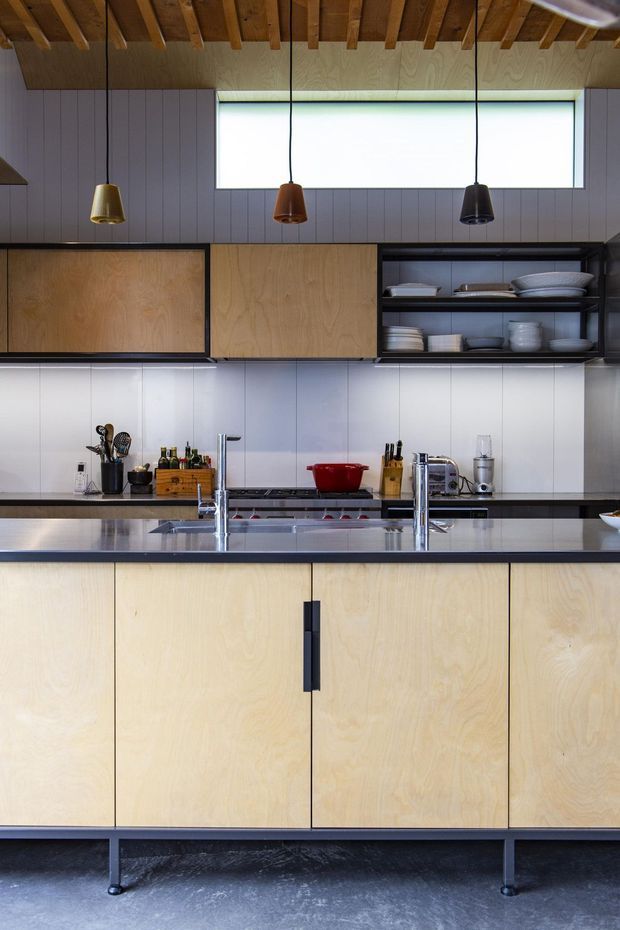 A contemporary galley-style kitchen with extensive pale timber plywood cabinetry, dark metal framing and handles, stainless-steel benchtops and sink, open shelving displaying crockery, and a white vertical-panel splashback. Three pendant lights hang beneath an exposed timber-ribbed ceiling, while a long horizontal window provides bright natural light.
