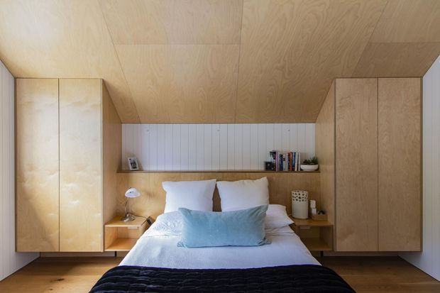 A compact, carefully integrated bedroom with a central bed framed by extensive light timber built-in cabinetry. The room features a timber-lined ceiling, pale vertical wall panelling behind the bed, open bedside shelves, and a recessed display shelf with books and small decor. Soft white bedding, a pale blue cushion, and a dark textured throw add restrained colour contrast.