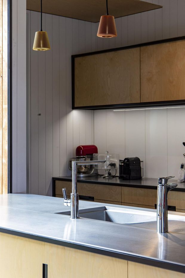 A contemporary kitchen viewed from the island, featuring a large pale stone island benchtop with an integrated double sink and polished chrome taps. Pale timber cabinetry with dark edging lines the back wall, paired with vertical white wall panelling and under-cabinet lighting. Two sculptural pendant lights hang from a timber-lined ceiling, while small countertop appliances sit in the background.