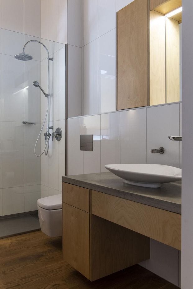 Contemporary bathroom with a frameless glass shower, chrome rain shower and handheld fixture, wall-hung toilet, vessel basin, and floating timber vanity with a dark stone benchtop. Large light tiles cover the walls, while warm timber flooring and matching timber cabinetry add contrast.