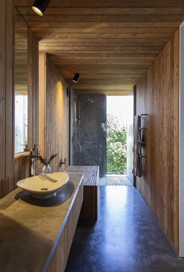 A narrow, elongated bathroom with warm timber wall and ceiling panelling, a concrete-look vanity with a white vessel basin, and a walk-in shower at the far end. The shower area opens toward an exterior deck and leafy outlook, creating a bright indoor-outdoor connection. Dark polished flooring, black directional ceiling lights, and a timber slatted bench add contrast and texture.