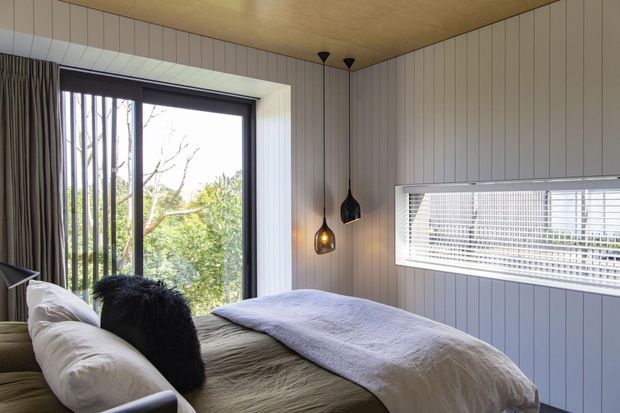 A cozy bedroom with a low-profile bed dressed in neutral linens, white vertical wall panelling, and a warm timber ceiling. Large windows with blinds and curtains bring in views of trees, while sculptural pendant lights provide soft ambient lighting. The room has a calm, natural, contemporary cabin-like atmosphere.