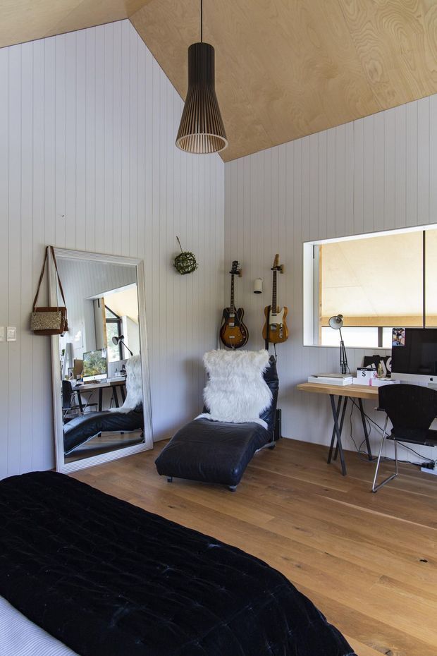 A compact, stylish bedroom with a home-office corner. The room features white vertical wall panelling, a warm plywood-lined sloped ceiling, timber flooring, a hanging sculptural pendant light, a wall mirror, guitars displayed on the wall, a black upholstered bed, and a black lounge chair with a white sheepskin throw. A desk with computer equipment sits beside a large interior window opening.