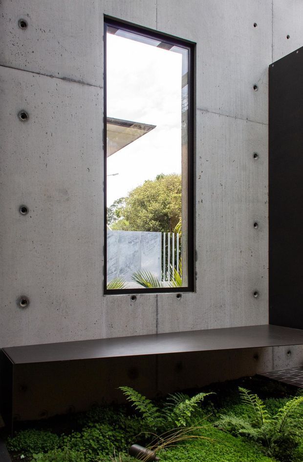 A narrow vertical window set into a raw, board-marked concrete wall, with dark metal framing and a deep built-in dark bench or ledge beneath it. An interior planting bed with ferns and groundcover occupies the foreground, while a fenced garden and trees are visible through the window. The composition is minimalist and architectural, emphasizing concrete, shadow, and greenery.