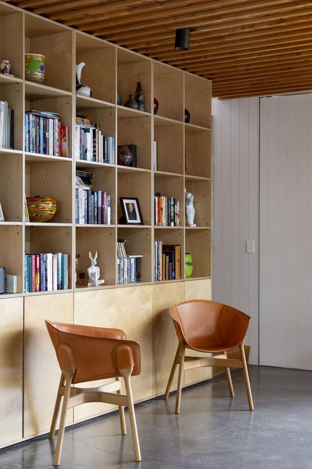 A refined reading nook or study area featuring a full-height built-in plywood bookshelf and lower cabinetry filled with books and decorative objects. Two cognac leather and timber lounge chairs sit on a polished concrete floor. The room has a warm exposed timber slat ceiling, pale vertical wall panelling, and a minimalist black ceiling spotlight.