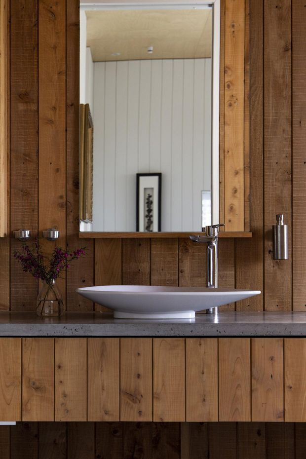 A warm rustic bathroom vanity with extensive vertical timber wall panelling and matching timber cabinetry. A white vessel sink sits on a grey stone benchtop beneath a large rectangular mirror, paired with a polished chrome wall-mounted tap and cylindrical wall sconce. Small decorative items add texture, while the reflected space shows pale vertical wall panelling and a timber ceiling.