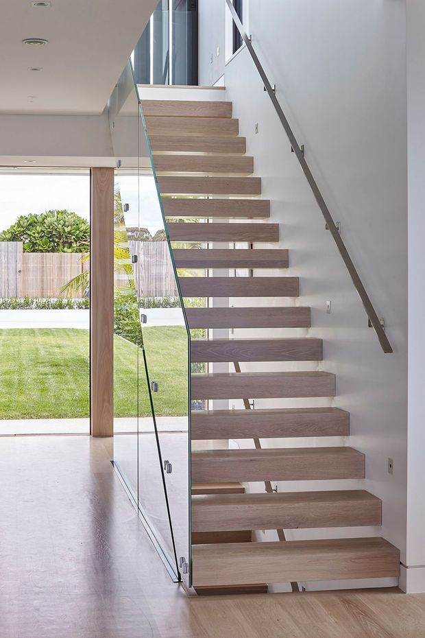 Bright contemporary interior focused on a floating staircase with thick timber treads, clear glass balustrade, and a slim dark metal handrail. The stair rises beside a white wall and opens onto a light-filled entry or circulation space, with timber flooring, recessed ceiling lights, and garden views through full-height glazing.