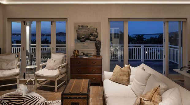 A warmly lit coastal living room with white upholstered seating, a timber chest, decorative pottery, a trunk-style coffee table and a zebra-pattern rug. Large glazed doors and windows open to a balcony with white railings and expansive waterfront views at dusk.