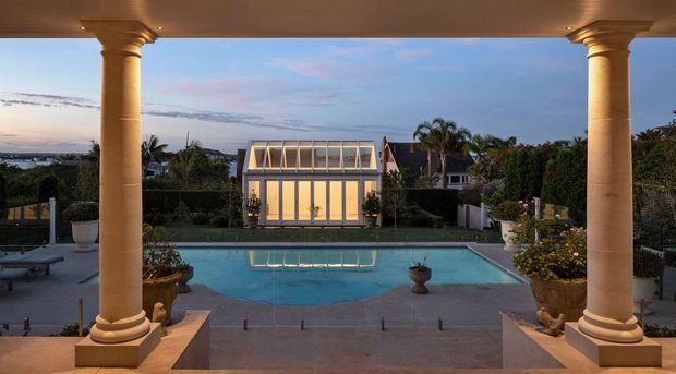 View from a covered terrace framed by two large classical columns, looking across a swimming pool toward a glazed pavilion or pool house. The landscaped garden includes hedges, palms, urn planters, and formal planting, with distant residential surroundings visible beyond.