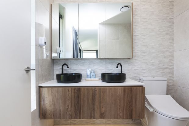 A contemporary bathroom vanity with two black vessel basins and matte black tapware set on a floating timber-look cabinet. A large mirrored cabinet spans the wall above the vanity, while pale patterned wall tiles, a white toilet, and a white door complete the compact, bright space.