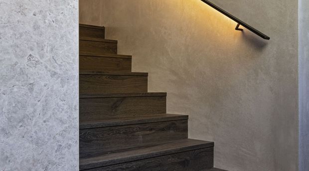 Close-up interior view of a modern stairway with dark timber stair treads and risers, a textured light-grey plaster wall, and a slim black handrail illuminated by concealed warm LED lighting.