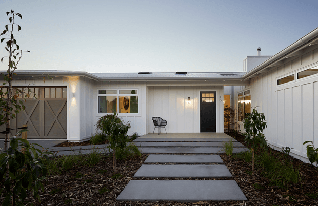Single-storey residential courtyard-style facade viewed from the front garden, with light vertical cladding, a low pitched metal roof, black-framed entry door, garage door, and broad concrete stepping slabs through landscaped planting. The exterior has a restrained contemporary farmhouse-like character with warm exterior wall lights.