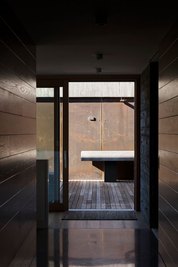 A dark, narrow residential entry hall leads through a glazed timber-framed doorway to an enclosed timber deck or courtyard. The hall has horizontal dark timber wall panelling, a polished dark floor, a plain dark ceiling with recessed cylindrical lights, and a contrasting rough dark stone wall at the right. Beyond the doorway is a weathered rust-toned metal privacy wall and a simple projecting bench or counter.