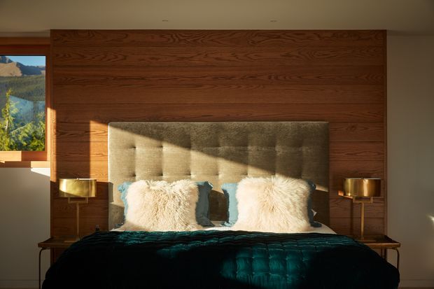 A warmly lit bedroom centered on a bed with a tall upholstered grey headboard, cream textured cushions, and a dark teal quilt. The bed is set against a full-width dark timber feature wall with integrated bedside tables and matching brass-toned lamps. A window at left provides a view of a mountainous landscape and strong late-afternoon sunlight.