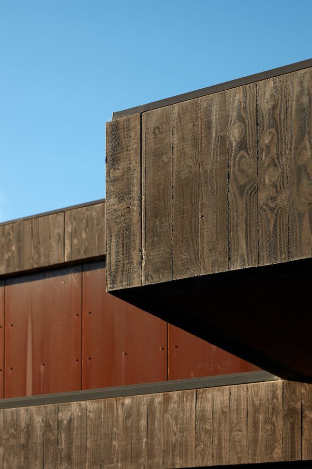 Close-up exterior architectural detail showing overlapping rectilinear volumes clad in weathered timber boards, with a rust-red metal wall panel beneath a projecting upper form. The composition emphasizes wood grain, dark shadow lines, crisp edges, and a clear blue sky, creating a contemporary, sculptural facade.
