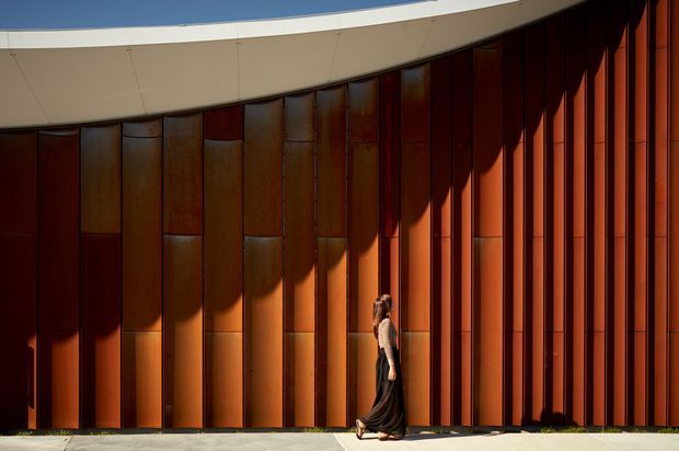 Exterior architectural detail featuring a dramatic curved facade clad in tall, vertically segmented rust-coloured metal panels. A smooth sweeping pale roof or canopy edge follows the curve above, while strong sunlight casts deep geometric shadows across the facade. A person standing at the base provides scale.