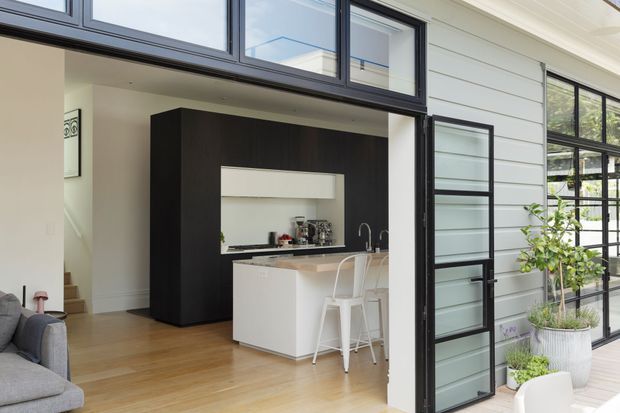 Bright contemporary open-plan kitchen viewed from an outdoor deck through a large black-framed glazed opening. The kitchen features a white island with bar seating, pale timber flooring, a dark wood-toned full-height cabinetry block, white horizontal wall cladding, and extensive black-framed glazing. A portion of the adjacent living area is visible inside, with potted plants and outdoor decking in the foreground.