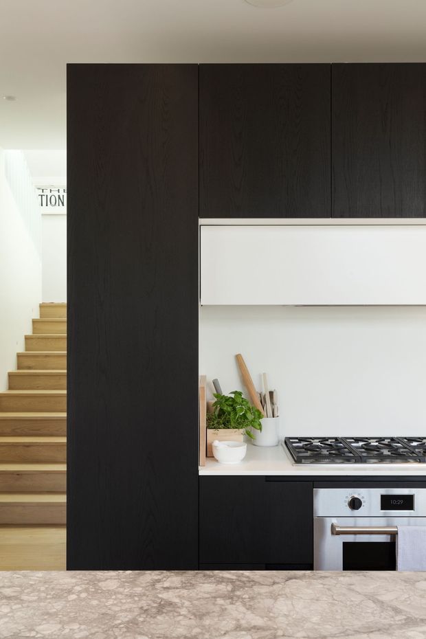 A compact contemporary kitchen with dark timber-grain cabinetry, white upper cabinetry and splashback, a stainless-steel gas cooktop and oven, and a pale veined stone island benchtop in the foreground. A timber staircase is visible beside the kitchen, with open shelving and small countertop items adding warmth.