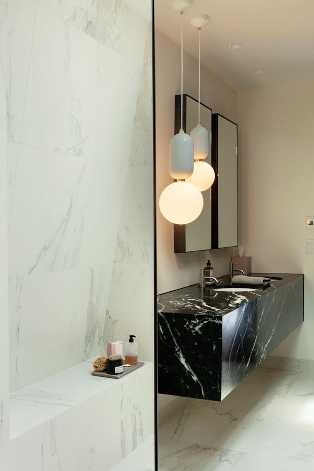 Contemporary bathroom featuring large white marble-look wall and floor slabs, a black veined stone floating vanity with integrated basin, mirrored wall cabinets, chrome tapware, and sculptural globe pendant lights. A partially visible marble-clad shower or bath zone is separated by a clear glass panel.