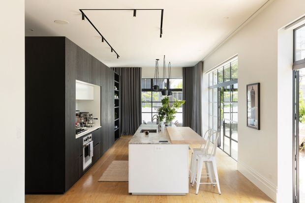 Bright contemporary kitchen with a long white island, integrated dark timber cabinetry, built-in appliances, pendant lights, timber flooring, and extensive black-framed glazing opening toward an outdoor area. A dining or living space is visible beyond the kitchen.