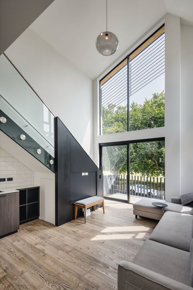 Bright contemporary double-height living room with a mezzanine-style stair and kitchen edge. The space features floor-to-ceiling black-framed glazing with horizontal blinds, white walls, pale timber flooring, a dark built-in stair wall, a grey sectional sofa, and a suspended spherical glass pendant. A glazed balustrade and open-riser stair contribute to the airy vertical volume.
