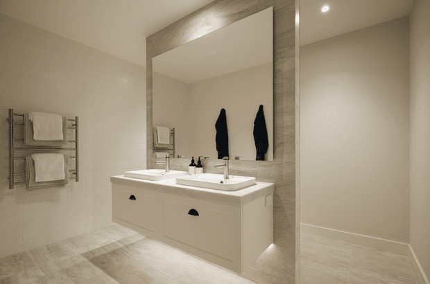 Contemporary double-vanity bathroom with a large frameless mirror set into a grey stone-look surround. The room features white subway wall tiles, pale large-format floor tiles, a wall-mounted white vanity with dark pulls, twin basins, chrome tapware, recessed ceiling lighting, and a heated towel rail.