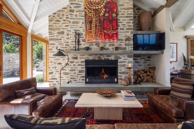 A warm rustic living room centred on a substantial rough-stone fireplace wall with a built-in fire, timber mantel, mounted television and stacked firewood. Leather sofas and armchairs surround a pale rectangular coffee table over a richly patterned rug. Large timber-framed windows and glazed doors open the room to leafy surroundings, while a white-painted vaulted ceiling with exposed timber beams adds architectural character.