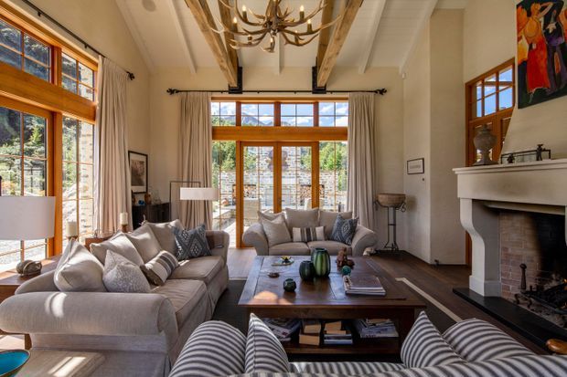 A bright, spacious living room with high ceilings, exposed timber beams, large timber-framed windows and glazed doors, and a traditional fireplace with a white mantel. Neutral upholstered sofas surround a substantial wooden coffee table, while an antler-style chandelier and artwork add character. The room opens visually to a landscaped exterior through extensive glazing.