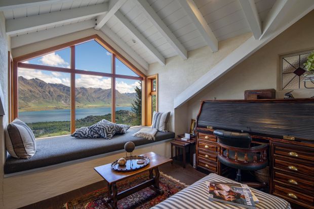 A cosy living room with a built-in window seat overlooking a dramatic alpine lake and mountain landscape. The room features exposed white-painted roof beams and sloping ceilings, warm timber flooring, a large upright piano, patterned rug, coffee table, and layered upholstered seating.